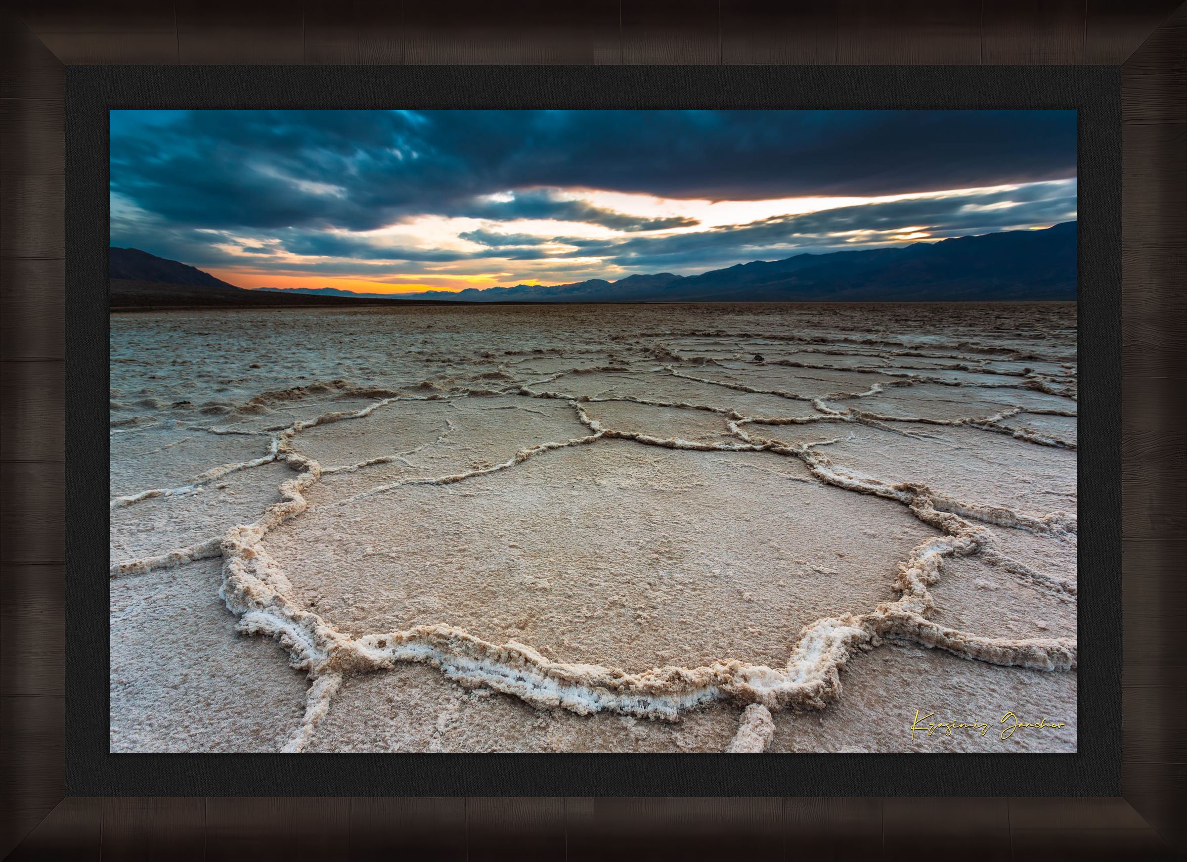 Expansive desert salt flat of Badwater Basin beneath a sky filled with storm clouds and setting sun in Death Valley. #Finish_Roma Dark Ash Frame & Dark Liner