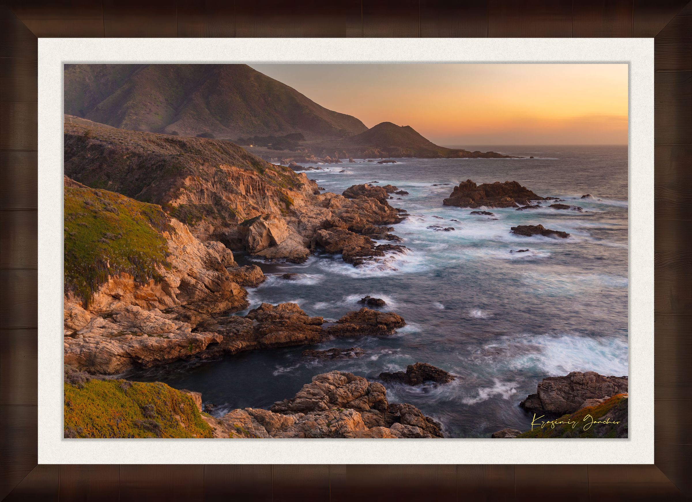 Rugged coastline of Big Sur featuring dark boulder formations, green slopes, ocean waves, and soft sunset glow across land and sea. #Finish_Roma Cigar Leaf Frame & Bright Liner