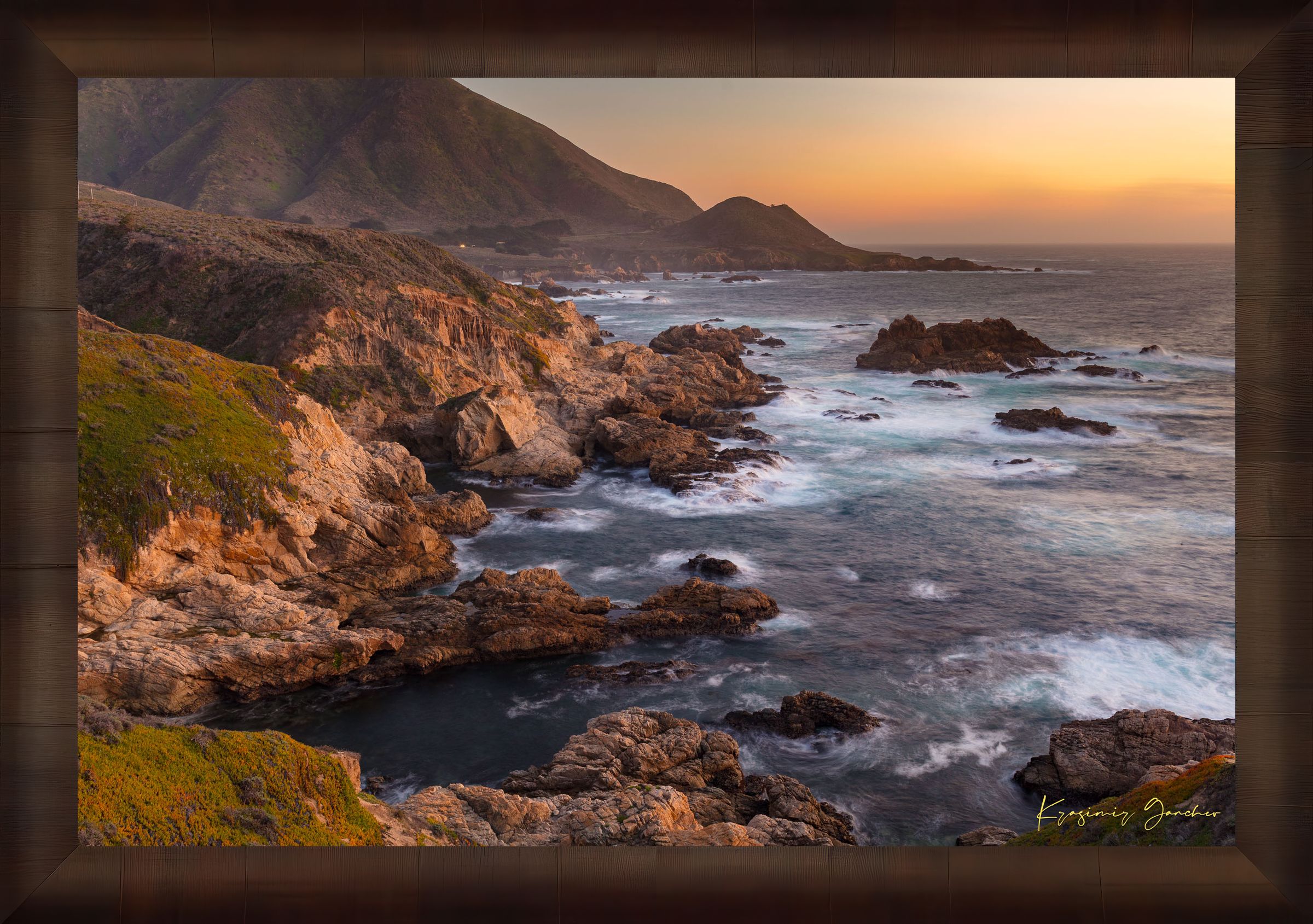 Rugged coastline of Big Sur featuring dark boulder formations, green slopes, ocean waves, and soft sunset glow across land and sea. #Finish_Roma Cigar Leaf Frame