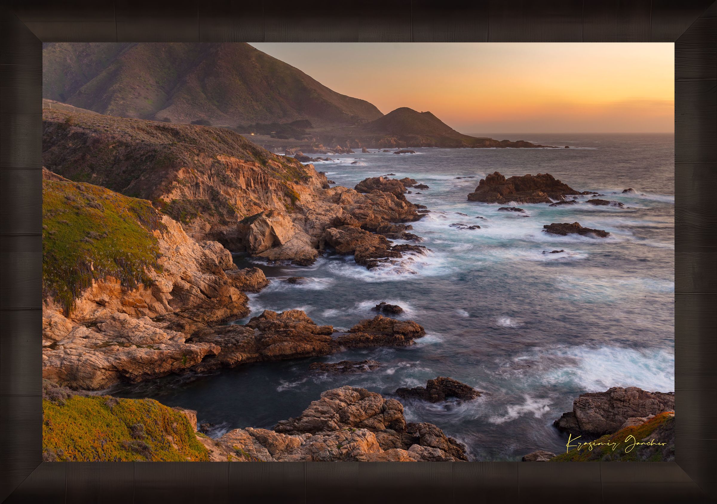 Rugged coastline of Big Sur featuring dark boulder formations, green slopes, ocean waves, and soft sunset glow across land and sea. #Finish_Roma Dark Ash Frame