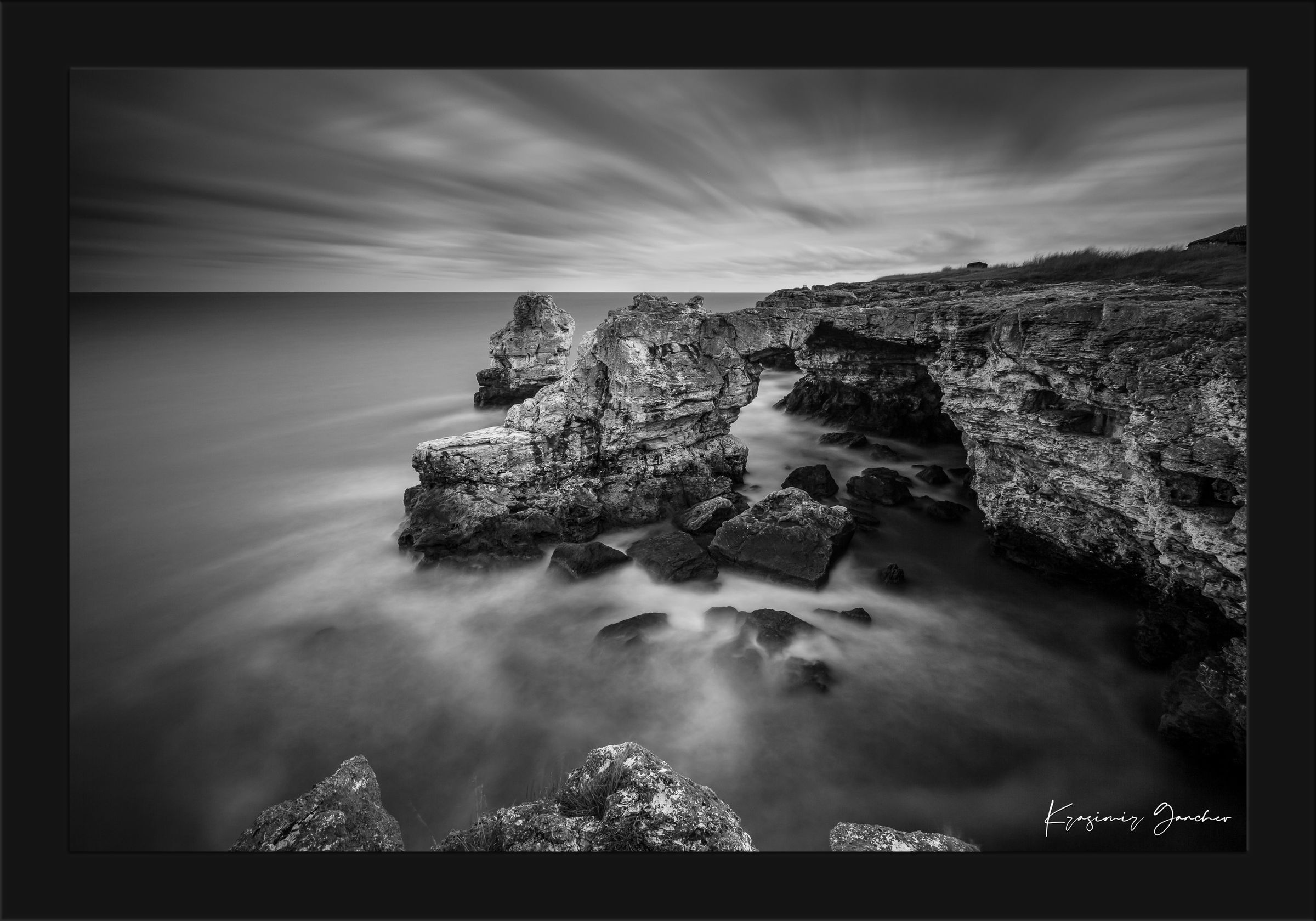 A stone arch formation near coastal cliffs in monochrome, with waves crashing against it. #Finish_Roma Satin Black Frame