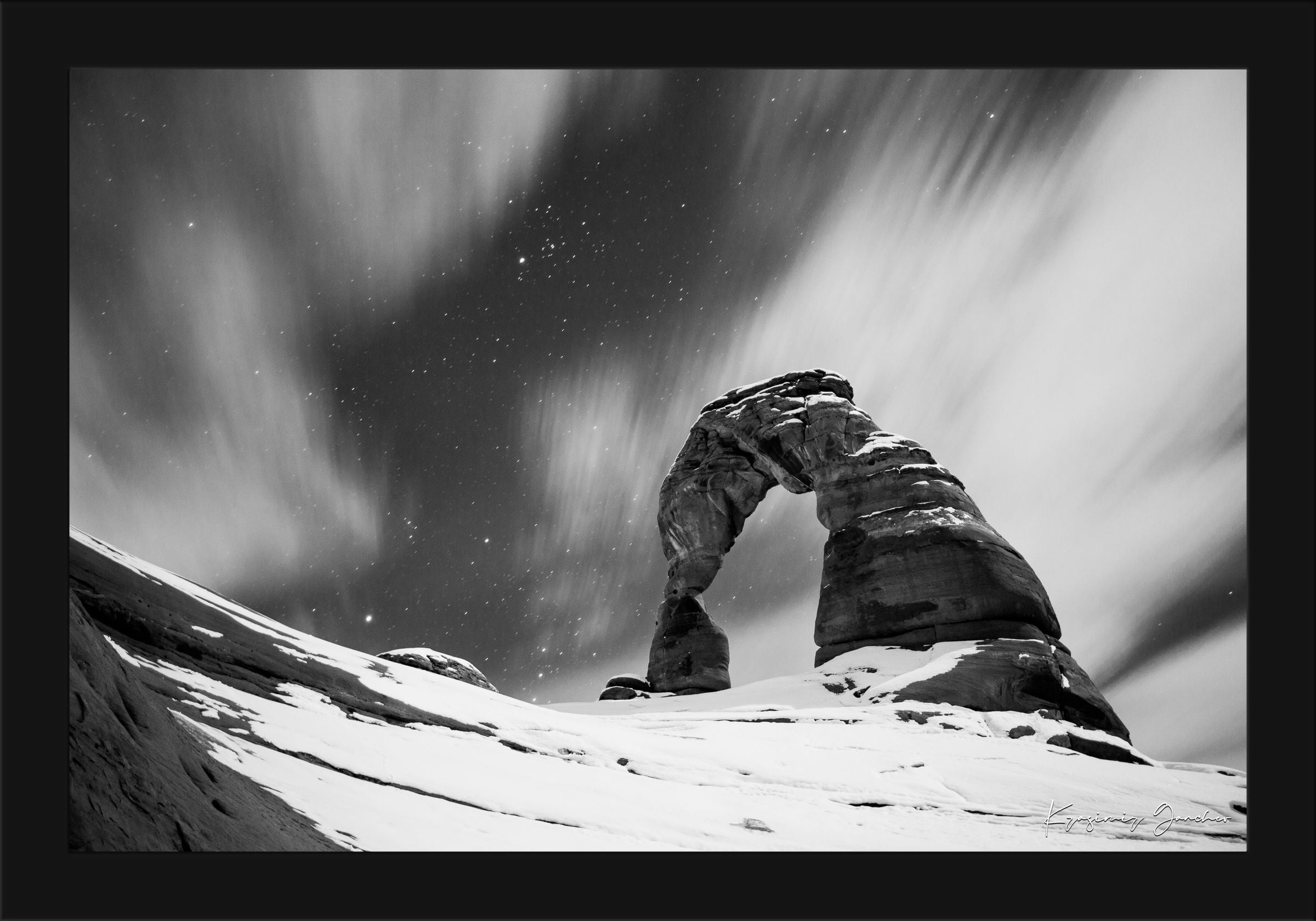 Silhouette of Delicate Arch against a star-filled night sky in Utah's Arches National Park, featuring cloud trails and snow cover. #Finish_Roma Satin Black Frame