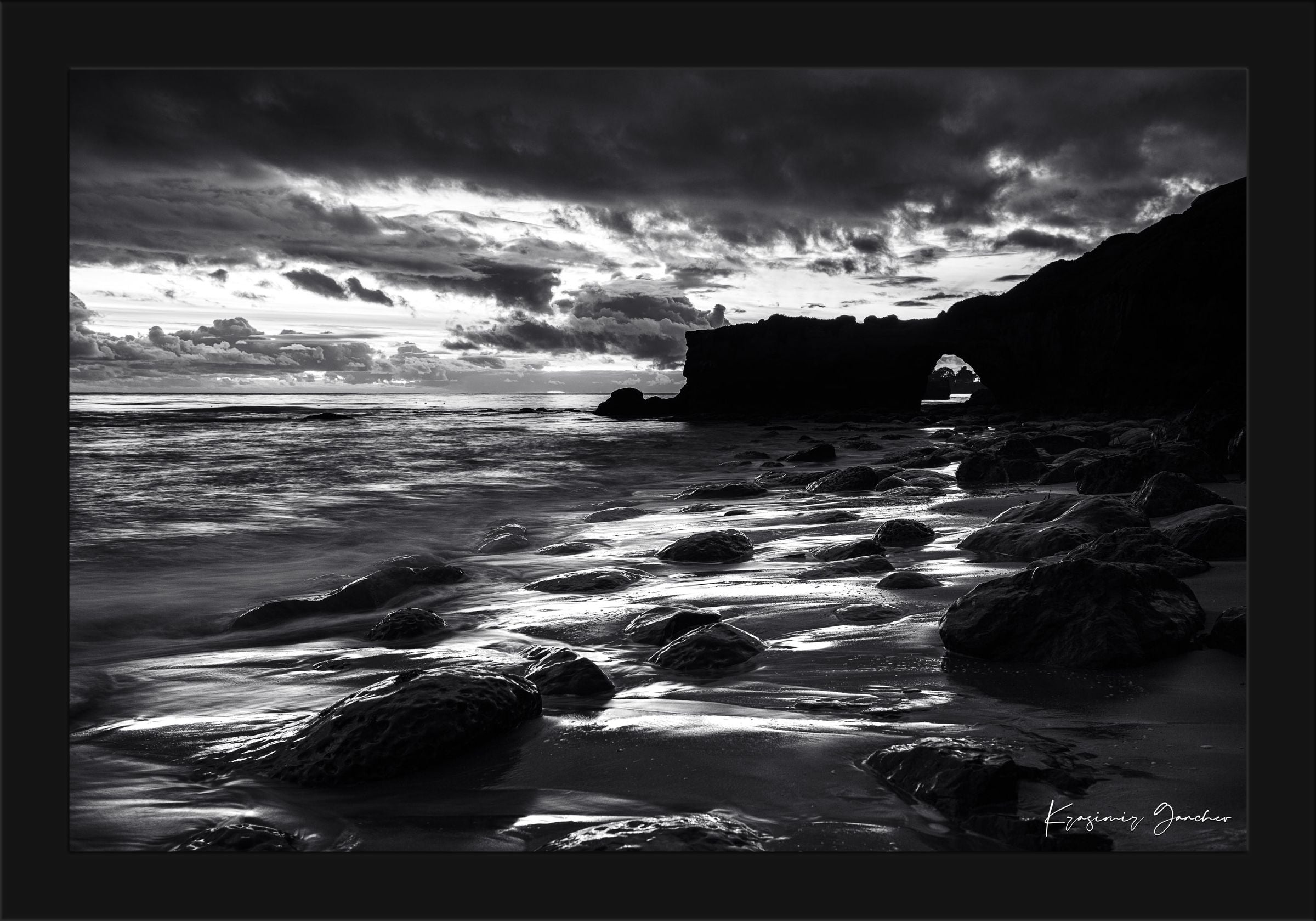Black-and-white photograph showcasing a rock arch at Santa Cruz’s coastline during sunset with flowing waves. #Finish_Roma Satin Black Frame