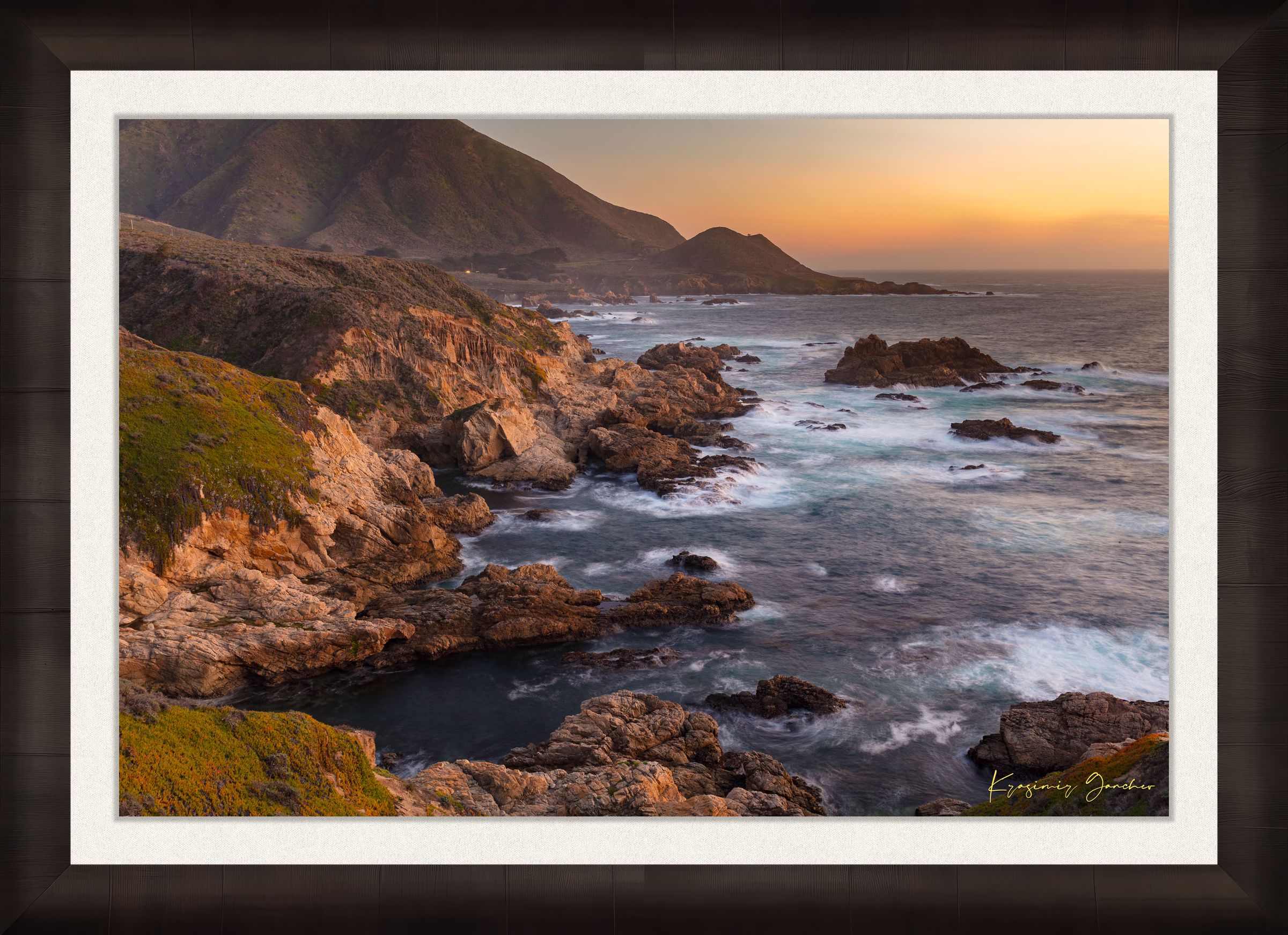 Rugged coastline of Big Sur featuring dark boulder formations, green slopes, ocean waves, and soft sunset glow across land and sea. #Finish_Roma Dark Ash Frame & Bright Liner