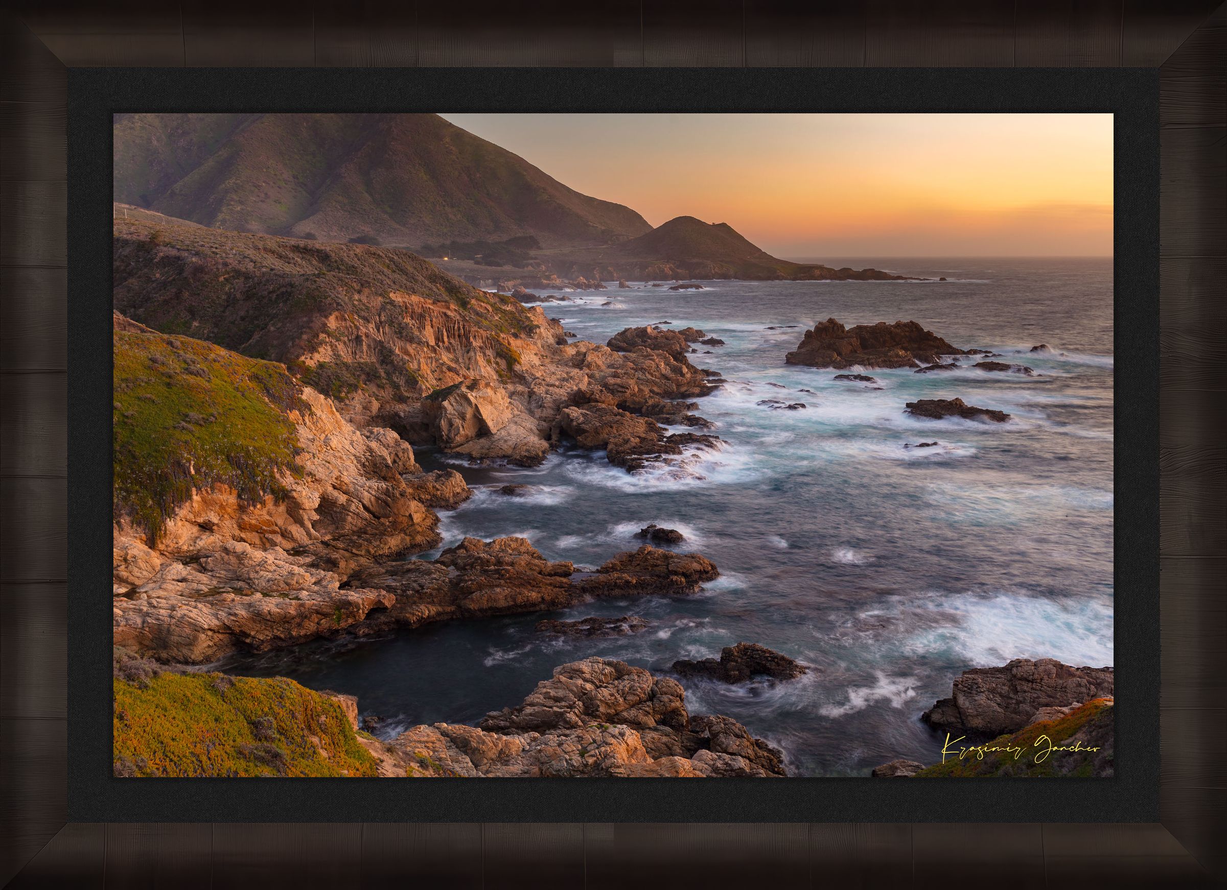 Rugged coastline of Big Sur featuring dark boulder formations, green slopes, ocean waves, and soft sunset glow across land and sea. #Finish_Roma Dark Ash Frame & Dark Liner
