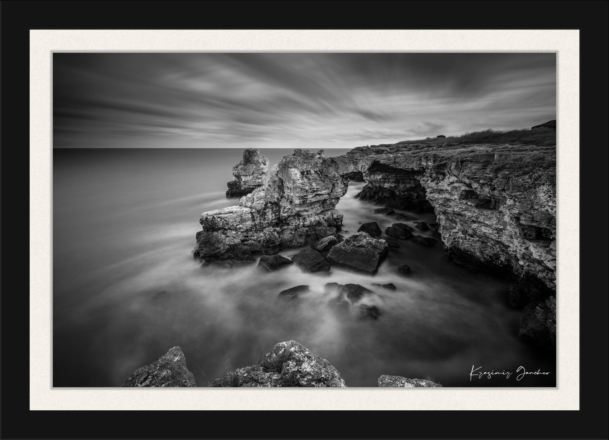 A stone arch formation near coastal cliffs in monochrome, with waves crashing against it. #Finish_Roma Satin Black Frame & Bright Liner