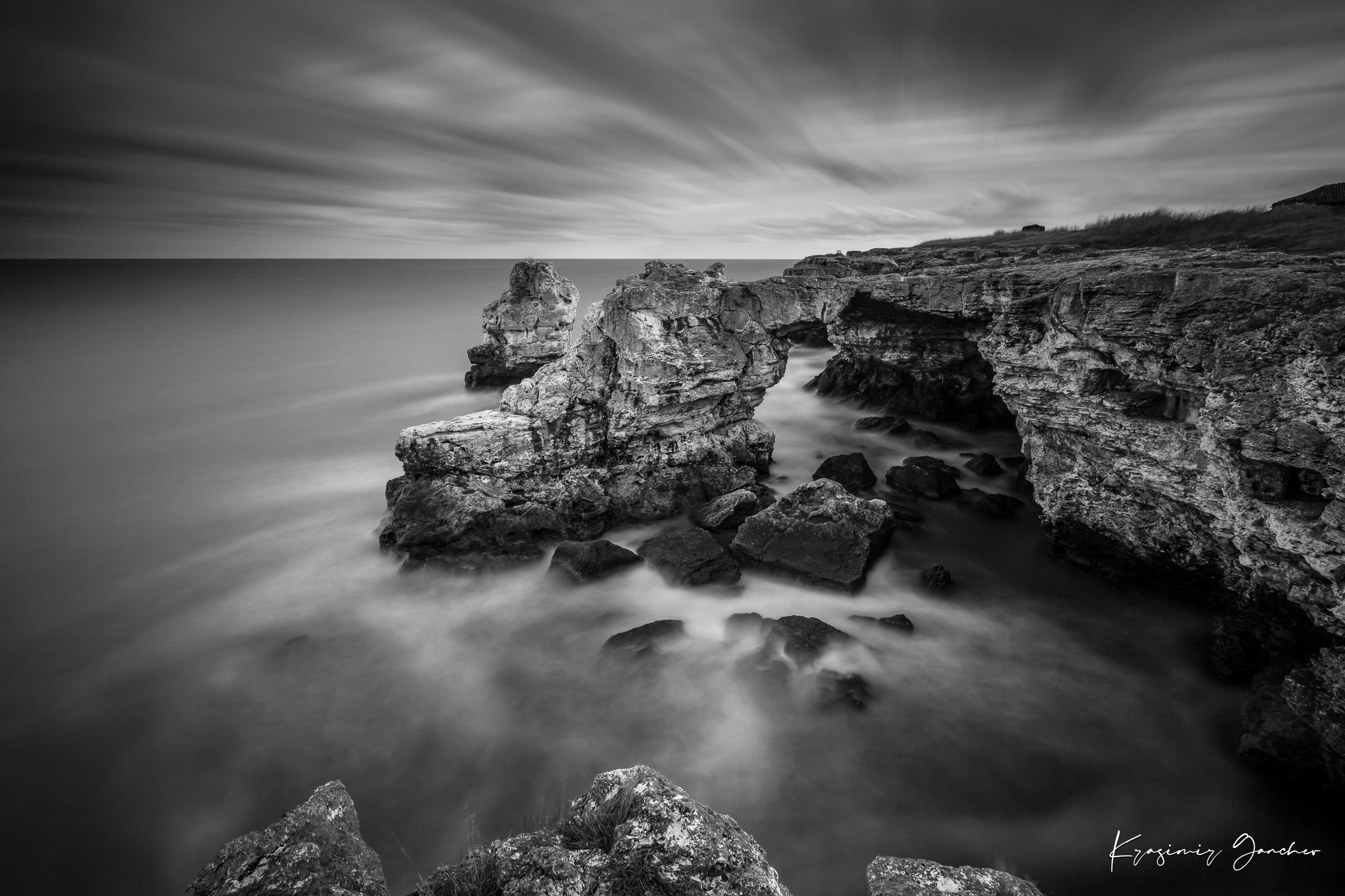 A stone arch formation near coastal cliffs in monochrome, with waves crashing against it. #Finish_Acrylic Recess