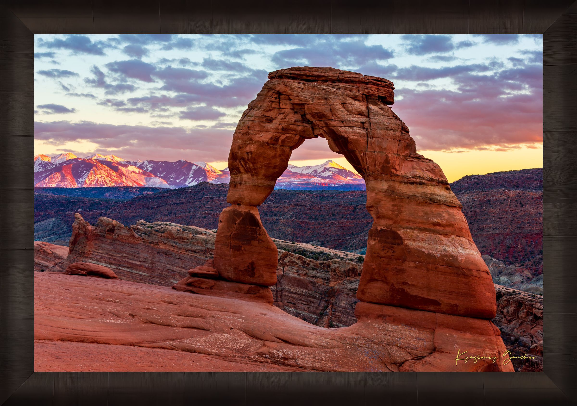 Natural arch of Delicate Arch in Arches National Park illuminated by sunset glow against a cloudy sky with vibrant reds and purples. #Finish_Roma Dark Ash Frame